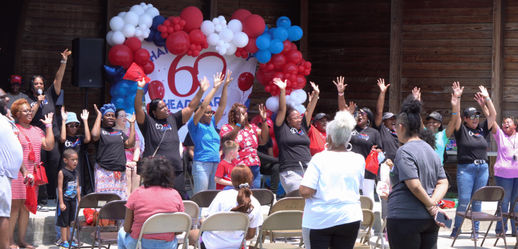 CASOMS Head Start staff, families, and community members celebrating on stage under red white and blue balloons at the 60th anniversary event at Edward Khayat Memorial Park in Moss Point